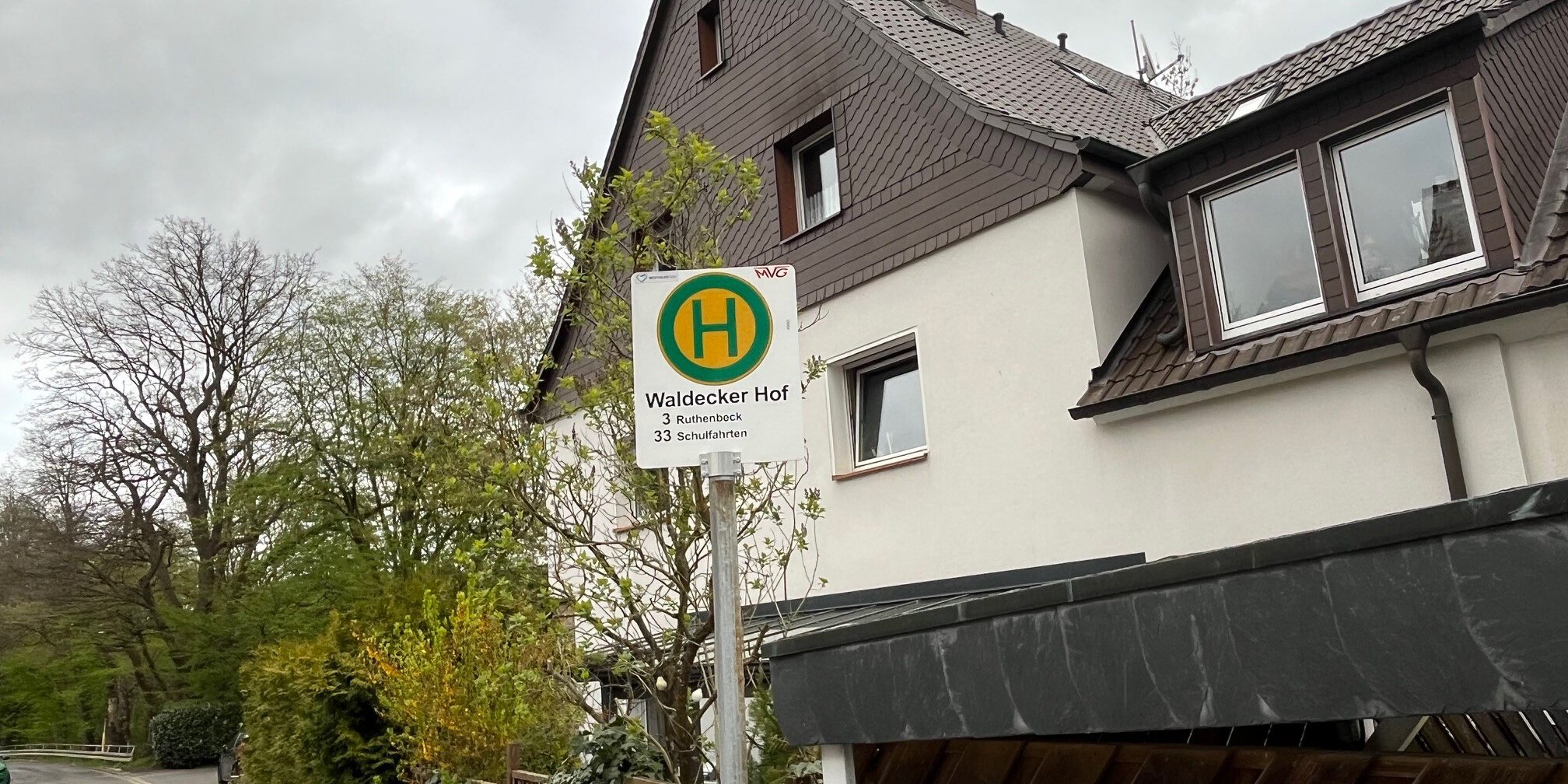 Ein Haltestellenschild mit der Aufschrift „Waldecker Hof“ steht vor einem Wohnhaus an einer ruhigen Straße, umgeben von Bäumen und Hecken unter grauem Himmel.