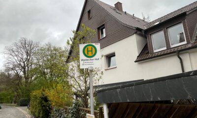 Ein Haltestellenschild mit der Aufschrift „Waldecker Hof“ steht vor einem Wohnhaus an einer ruhigen Straße, umgeben von Bäumen und Hecken unter grauem Himmel.