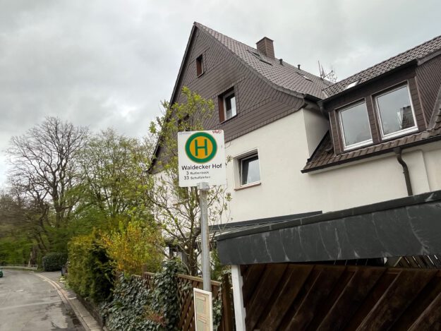 Ein Haltestellenschild mit der Aufschrift „Waldecker Hof“ steht vor einem Wohnhaus an einer ruhigen Straße, umgeben von Bäumen und Hecken unter grauem Himmel.