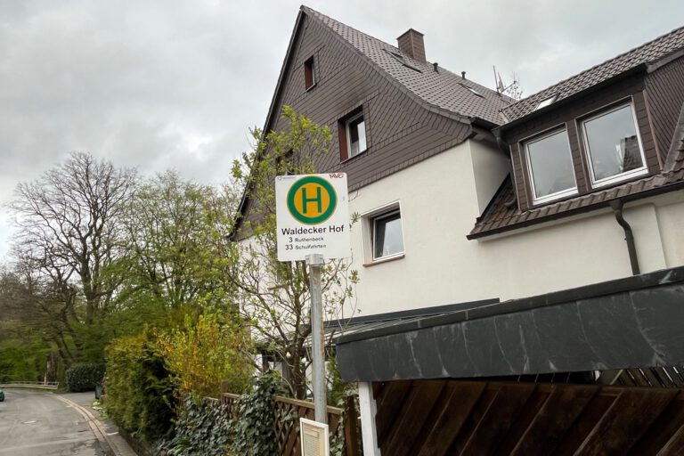 Ein Haltestellenschild mit der Aufschrift „Waldecker Hof“ steht vor einem Wohnhaus an einer ruhigen Straße, umgeben von Bäumen und Hecken unter grauem Himmel.