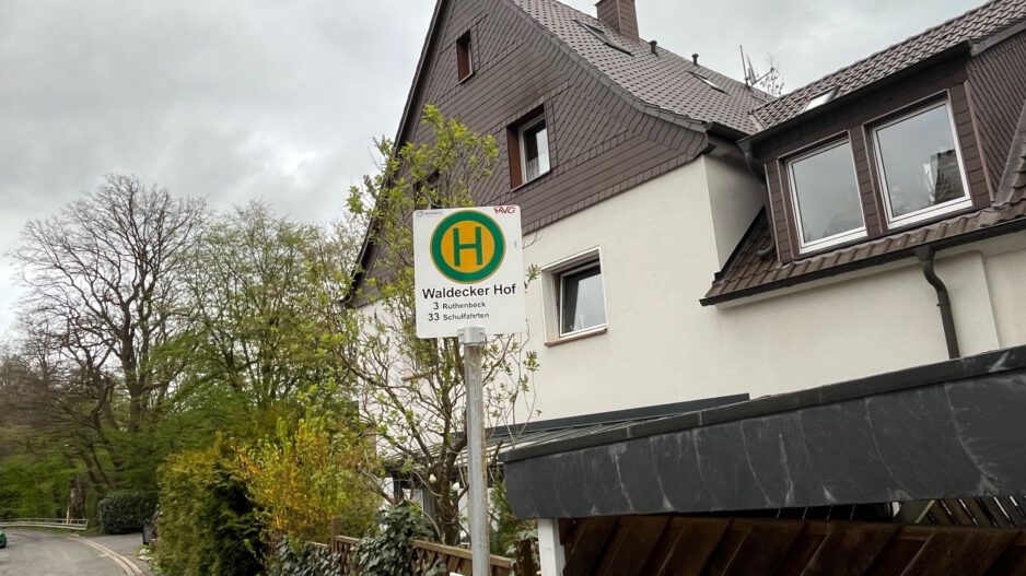 Ein Haltestellenschild mit der Aufschrift „Waldecker Hof“ steht vor einem Wohnhaus an einer ruhigen Straße, umgeben von Bäumen und Hecken unter grauem Himmel.