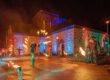 Vor einem historischen Backstein- und Natursteingebäude findet bei Nacht eine spektakuläre Feuershow statt. Mehrere Künstlerinnen und Künstler schwingen brennende Requisiten und erzeugen leuchtende Feuerkreise. Zahlreiche Zuschauer verfolgen die Aufführung, während das Gebäude mit farbigen Lichtakzenten in Blau, Rot und Grün illuminiert wird. Die Szene vermittelt die besondere Atmosphäre einer Kultur- oder Festveranstaltung unter freiem Himmel.
