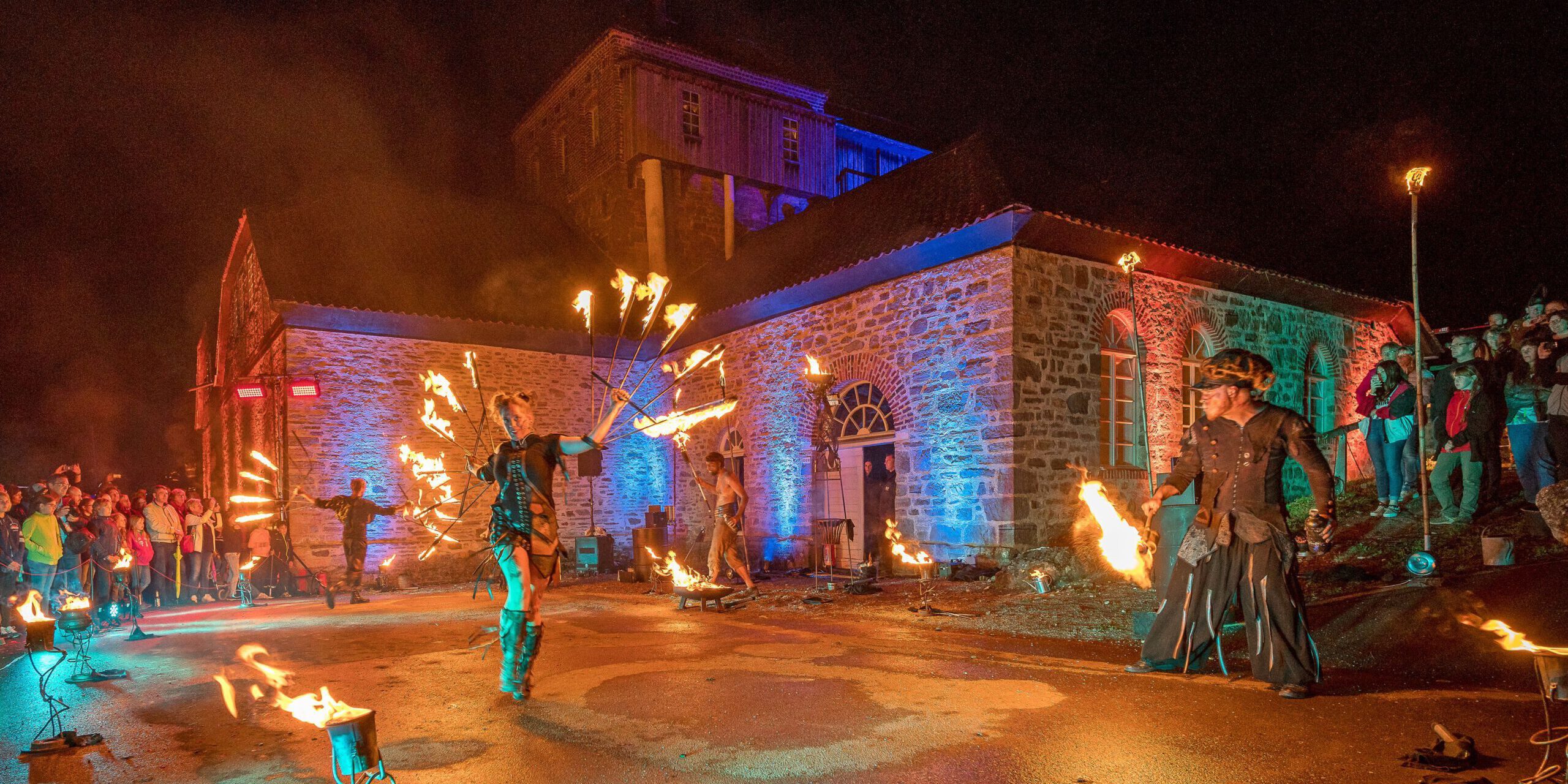 Vor einem historischen Backstein- und Natursteingebäude findet bei Nacht eine spektakuläre Feuershow statt. Mehrere Künstlerinnen und Künstler schwingen brennende Requisiten und erzeugen leuchtende Feuerkreise. Zahlreiche Zuschauer verfolgen die Aufführung, während das Gebäude mit farbigen Lichtakzenten in Blau, Rot und Grün illuminiert wird. Die Szene vermittelt die besondere Atmosphäre einer Kultur- oder Festveranstaltung unter freiem Himmel.