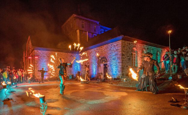 Vor einem historischen Backstein- und Natursteingebäude findet bei Nacht eine spektakuläre Feuershow statt. Mehrere Künstlerinnen und Künstler schwingen brennende Requisiten und erzeugen leuchtende Feuerkreise. Zahlreiche Zuschauer verfolgen die Aufführung, während das Gebäude mit farbigen Lichtakzenten in Blau, Rot und Grün illuminiert wird. Die Szene vermittelt die besondere Atmosphäre einer Kultur- oder Festveranstaltung unter freiem Himmel.