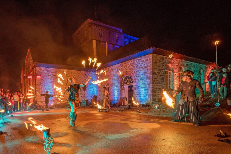 Foto: Stephan Sensen/Märkischer Kreis Vor einem historischen Backstein- und Natursteingebäude findet bei Nacht eine spektakuläre Feuershow statt. Mehrere Künstlerinnen und Künstler schwingen brennende Requisiten und erzeugen leuchtende Feuerkreise. Zahlreiche Zuschauer verfolgen die Aufführung, während das Gebäude mit farbigen Lichtakzenten in Blau, Rot und Grün illuminiert wird. Die Szene vermittelt die besondere Atmosphäre einer Kultur- oder Festveranstaltung unter freiem Himmel.