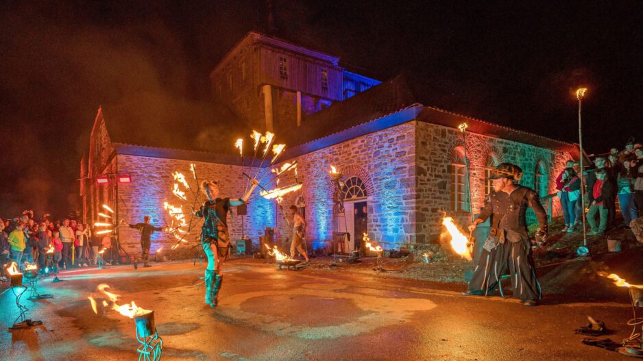 Vor einem historischen Backstein- und Natursteingebäude findet bei Nacht eine spektakuläre Feuershow statt. Mehrere Künstlerinnen und Künstler schwingen brennende Requisiten und erzeugen leuchtende Feuerkreise. Zahlreiche Zuschauer verfolgen die Aufführung, während das Gebäude mit farbigen Lichtakzenten in Blau, Rot und Grün illuminiert wird. Die Szene vermittelt die besondere Atmosphäre einer Kultur- oder Festveranstaltung unter freiem Himmel.