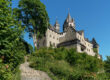 Die Burg Altena erhebt sich auf einem bewaldeten Hügel. Mehrere Türme und Gebäude prägen das Erscheinungsbild der Festung, die über einen steilen Treppenweg durch üppiges Grün erreichbar ist. Über der Anlage spannt sich ein wolkenloser blauer Himmel.