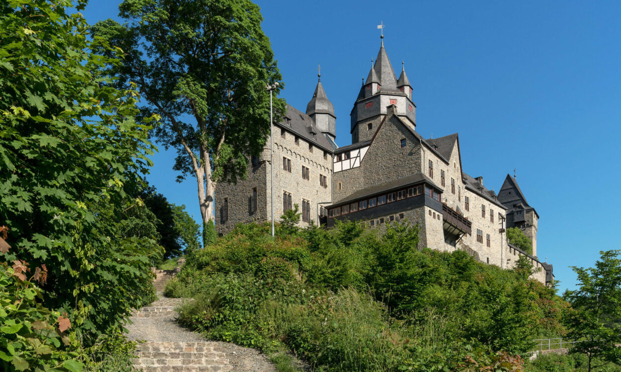 Die Burg Altena erhebt sich auf einem bewaldeten Hügel. Mehrere Türme und Gebäude prägen das Erscheinungsbild der Festung, die über einen steilen Treppenweg durch üppiges Grün erreichbar ist. Über der Anlage spannt sich ein wolkenloser blauer Himmel.