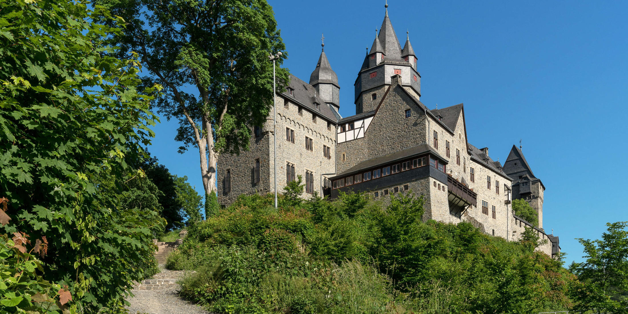 Die Burg Altena erhebt sich auf einem bewaldeten Hügel. Mehrere Türme und Gebäude prägen das Erscheinungsbild der Festung, die über einen steilen Treppenweg durch üppiges Grün erreichbar ist. Über der Anlage spannt sich ein wolkenloser blauer Himmel.