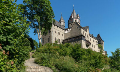Die Burg Altena erhebt sich auf einem bewaldeten Hügel. Mehrere Türme und Gebäude prägen das Erscheinungsbild der Festung, die über einen steilen Treppenweg durch üppiges Grün erreichbar ist. Über der Anlage spannt sich ein wolkenloser blauer Himmel.