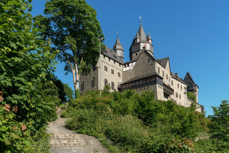 Die Burg Altena erhebt sich auf einem bewaldeten Hügel. Mehrere Türme und Gebäude prägen das Erscheinungsbild der Festung, die über einen steilen Treppenweg durch üppiges Grün erreichbar ist. Über der Anlage spannt sich ein wolkenloser blauer Himmel.