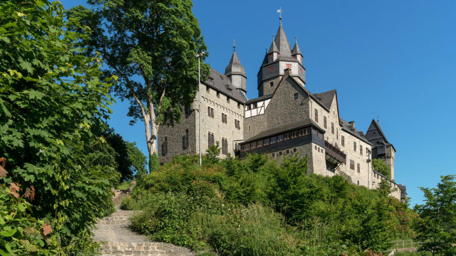 Die Burg Altena erhebt sich auf einem bewaldeten Hügel. Mehrere Türme und Gebäude prägen das Erscheinungsbild der Festung, die über einen steilen Treppenweg durch üppiges Grün erreichbar ist. Über der Anlage spannt sich ein wolkenloser blauer Himmel.