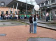 Auf dem Schulhof einer Schule haben sich zahlreiche Kinder zu einer gemeinsamen Veranstaltung versammelt. Im Vordergrund stehen zwei Schülerinnen mit dem Rücken zur Kamera und halten einen großen weißen Ballon. Ihnen gegenüber spricht eine Frau vor der versammelten Schulgemeinschaft. Die Kinder stehen in Gruppen vor dem Schulgebäude mit Glasdach und überdachtem Eingangsbereich. Der Boden ist noch feucht, was auf einen vorherigen Regenschauer hindeutet. Die Szene vermittelt den Eindruck einer Gedenk- oder Gemeinschaftsaktion im Freien.