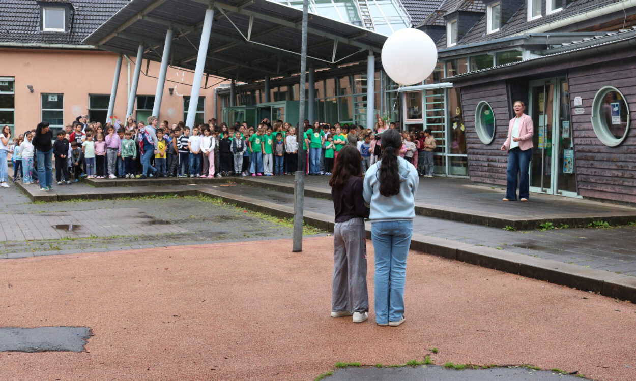 Auf dem Schulhof einer Schule haben sich zahlreiche Kinder zu einer gemeinsamen Veranstaltung versammelt. Im Vordergrund stehen zwei Schülerinnen mit dem Rücken zur Kamera und halten einen großen weißen Ballon. Ihnen gegenüber spricht eine Frau vor der versammelten Schulgemeinschaft. Die Kinder stehen in Gruppen vor dem Schulgebäude mit Glasdach und überdachtem Eingangsbereich. Der Boden ist noch feucht, was auf einen vorherigen Regenschauer hindeutet. Die Szene vermittelt den Eindruck einer Gedenk- oder Gemeinschaftsaktion im Freien.