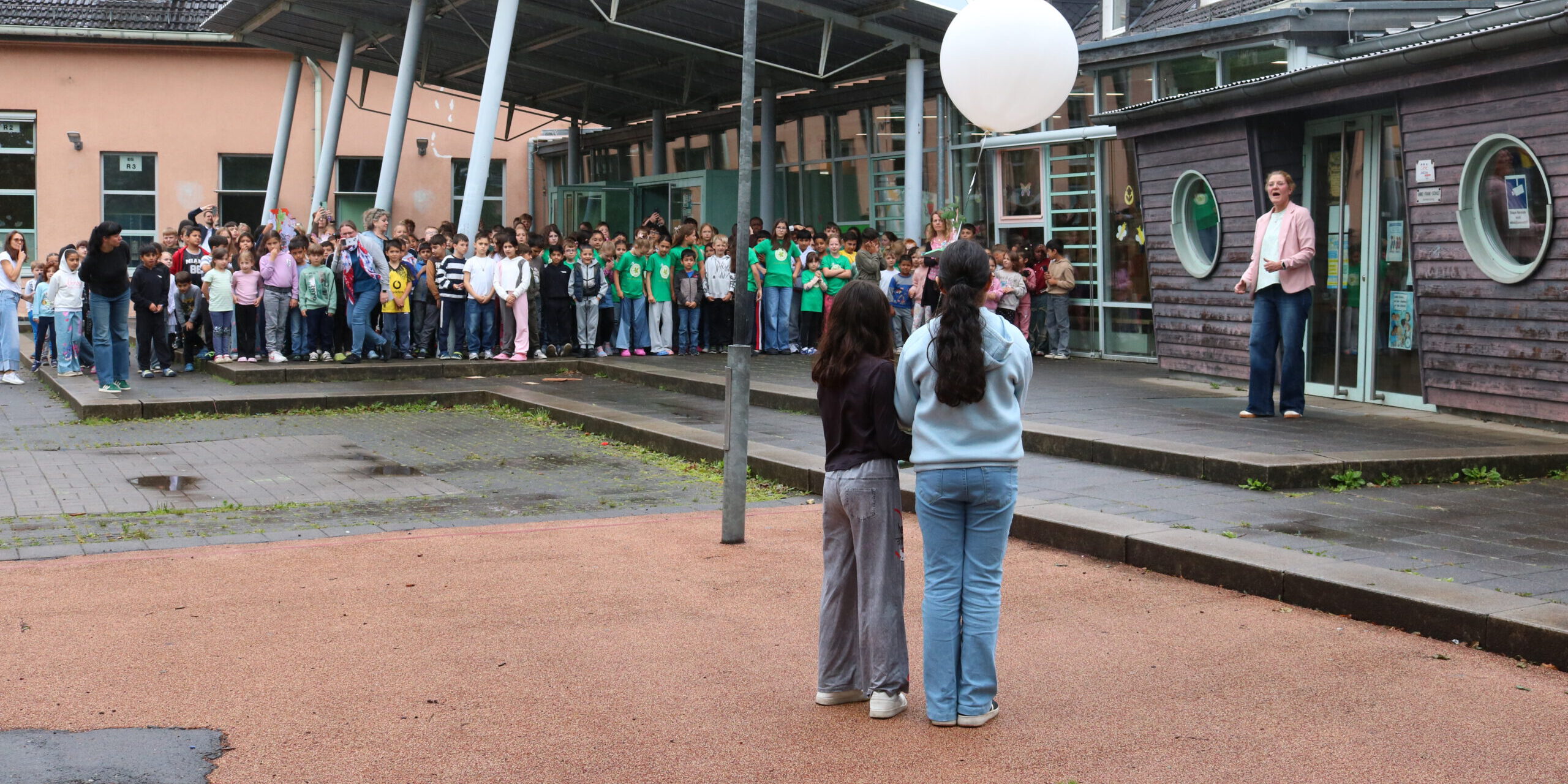 Auf dem Schulhof einer Schule haben sich zahlreiche Kinder zu einer gemeinsamen Veranstaltung versammelt. Im Vordergrund stehen zwei Schülerinnen mit dem Rücken zur Kamera und halten einen großen weißen Ballon. Ihnen gegenüber spricht eine Frau vor der versammelten Schulgemeinschaft. Die Kinder stehen in Gruppen vor dem Schulgebäude mit Glasdach und überdachtem Eingangsbereich. Der Boden ist noch feucht, was auf einen vorherigen Regenschauer hindeutet. Die Szene vermittelt den Eindruck einer Gedenk- oder Gemeinschaftsaktion im Freien.