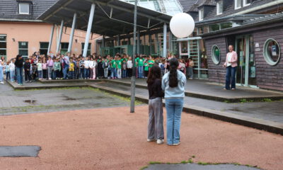 Auf dem Schulhof einer Schule haben sich zahlreiche Kinder zu einer gemeinsamen Veranstaltung versammelt. Im Vordergrund stehen zwei Schülerinnen mit dem Rücken zur Kamera und halten einen großen weißen Ballon. Ihnen gegenüber spricht eine Frau vor der versammelten Schulgemeinschaft. Die Kinder stehen in Gruppen vor dem Schulgebäude mit Glasdach und überdachtem Eingangsbereich. Der Boden ist noch feucht, was auf einen vorherigen Regenschauer hindeutet. Die Szene vermittelt den Eindruck einer Gedenk- oder Gemeinschaftsaktion im Freien.