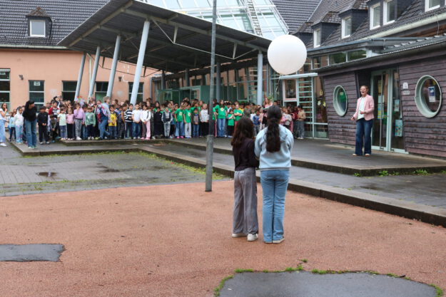 Auf dem Schulhof einer Schule haben sich zahlreiche Kinder zu einer gemeinsamen Veranstaltung versammelt. Im Vordergrund stehen zwei Schülerinnen mit dem Rücken zur Kamera und halten einen großen weißen Ballon. Ihnen gegenüber spricht eine Frau vor der versammelten Schulgemeinschaft. Die Kinder stehen in Gruppen vor dem Schulgebäude mit Glasdach und überdachtem Eingangsbereich. Der Boden ist noch feucht, was auf einen vorherigen Regenschauer hindeutet. Die Szene vermittelt den Eindruck einer Gedenk- oder Gemeinschaftsaktion im Freien.