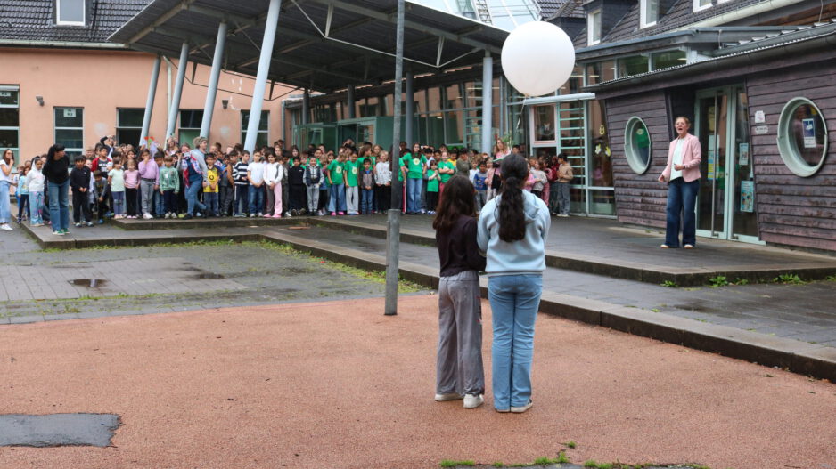 Auf dem Schulhof einer Schule haben sich zahlreiche Kinder zu einer gemeinsamen Veranstaltung versammelt. Im Vordergrund stehen zwei Schülerinnen mit dem Rücken zur Kamera und halten einen großen weißen Ballon. Ihnen gegenüber spricht eine Frau vor der versammelten Schulgemeinschaft. Die Kinder stehen in Gruppen vor dem Schulgebäude mit Glasdach und überdachtem Eingangsbereich. Der Boden ist noch feucht, was auf einen vorherigen Regenschauer hindeutet. Die Szene vermittelt den Eindruck einer Gedenk- oder Gemeinschaftsaktion im Freien.
