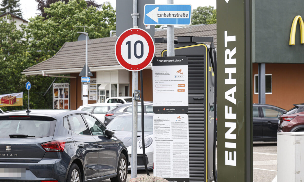 Hinweisschilder und geparkte Autos auf dem Parkplatz eines Schnellrestaurants. Im Vordergrund stehen ein Tempo-10-Schild, ein Einbahnstraßenschild sowie eine Informationstafel zur Parkdauerüberwachung. Rechts ist ein Teil des Restaurantgebäudes mit dem Schriftzug „Einfahrt“ zu sehen.