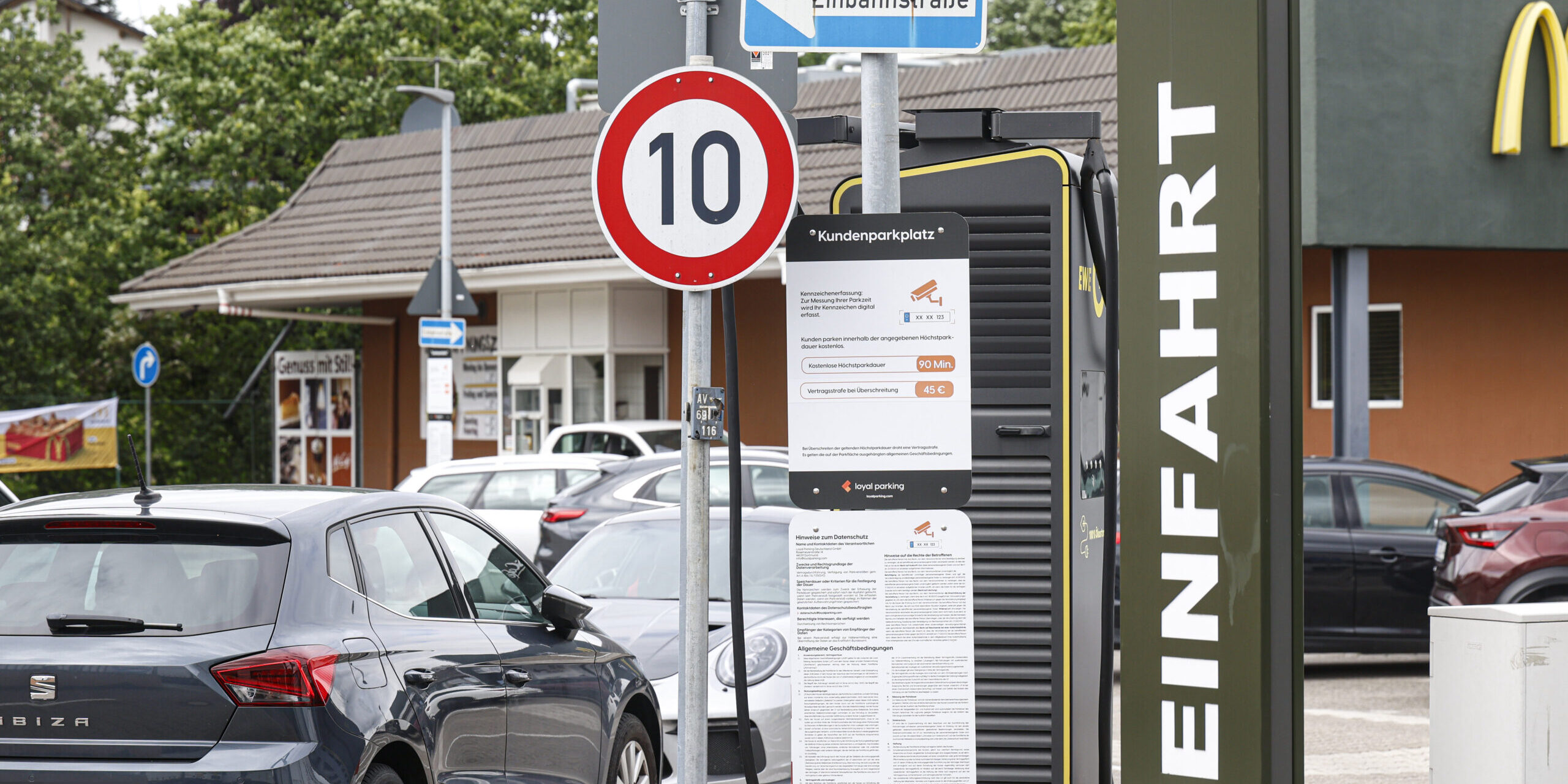 Hinweisschilder und geparkte Autos auf dem Parkplatz eines Schnellrestaurants. Im Vordergrund stehen ein Tempo-10-Schild, ein Einbahnstraßenschild sowie eine Informationstafel zur Parkdauerüberwachung. Rechts ist ein Teil des Restaurantgebäudes mit dem Schriftzug „Einfahrt“ zu sehen.