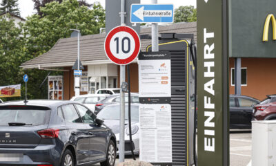 Hinweisschilder und geparkte Autos auf dem Parkplatz eines Schnellrestaurants. Im Vordergrund stehen ein Tempo-10-Schild, ein Einbahnstraßenschild sowie eine Informationstafel zur Parkdauerüberwachung. Rechts ist ein Teil des Restaurantgebäudes mit dem Schriftzug „Einfahrt“ zu sehen.