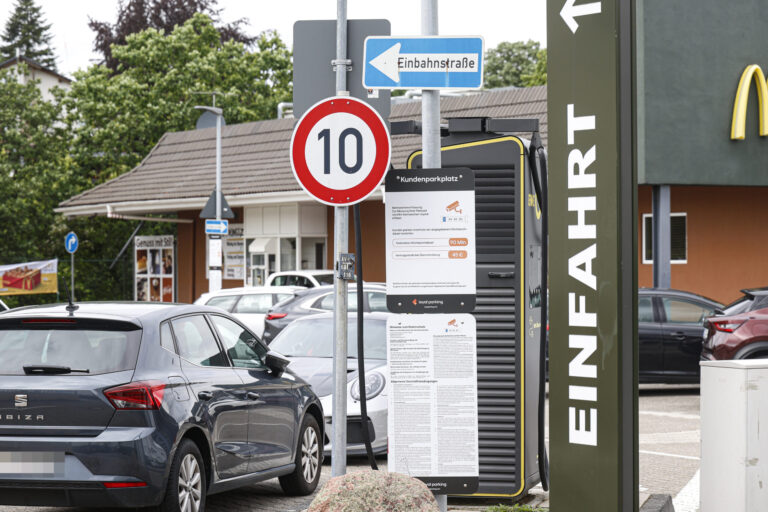 Hinweisschilder und geparkte Autos auf dem Parkplatz eines Schnellrestaurants. Im Vordergrund stehen ein Tempo-10-Schild, ein Einbahnstraßenschild sowie eine Informationstafel zur Parkdauerüberwachung. Rechts ist ein Teil des Restaurantgebäudes mit dem Schriftzug „Einfahrt“ zu sehen.