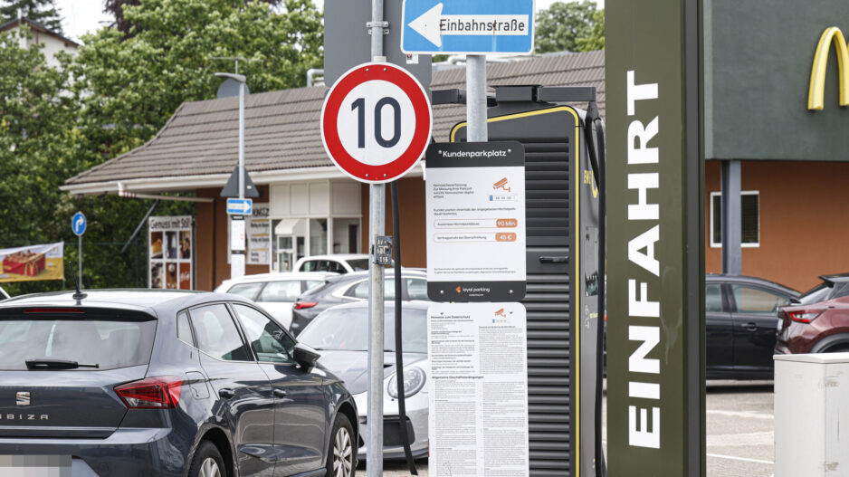 Hinweisschilder und geparkte Autos auf dem Parkplatz eines Schnellrestaurants. Im Vordergrund stehen ein Tempo-10-Schild, ein Einbahnstraßenschild sowie eine Informationstafel zur Parkdauerüberwachung. Rechts ist ein Teil des Restaurantgebäudes mit dem Schriftzug „Einfahrt“ zu sehen.