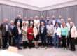 Gruppenfoto einer Ehrungsveranstaltung mit mehr als 20 Personen in einem Saal. Die Teilnehmenden stehen in mehreren Reihen vor einer grauen Vorhangwand. Im Vordergrund befindet sich eine Frau mit Bürgermeisterkette, umgeben von den geehrten Bürgerinnen und Bürgern verschiedener Altersgruppen. Links im Bild ist ein Klavier zu sehen.