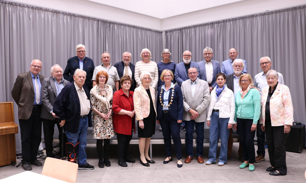 Gruppenfoto einer Ehrungsveranstaltung mit mehr als 20 Personen in einem Saal. Die Teilnehmenden stehen in mehreren Reihen vor einer grauen Vorhangwand. Im Vordergrund befindet sich eine Frau mit Bürgermeisterkette, umgeben von den geehrten Bürgerinnen und Bürgern verschiedener Altersgruppen. Links im Bild ist ein Klavier zu sehen.