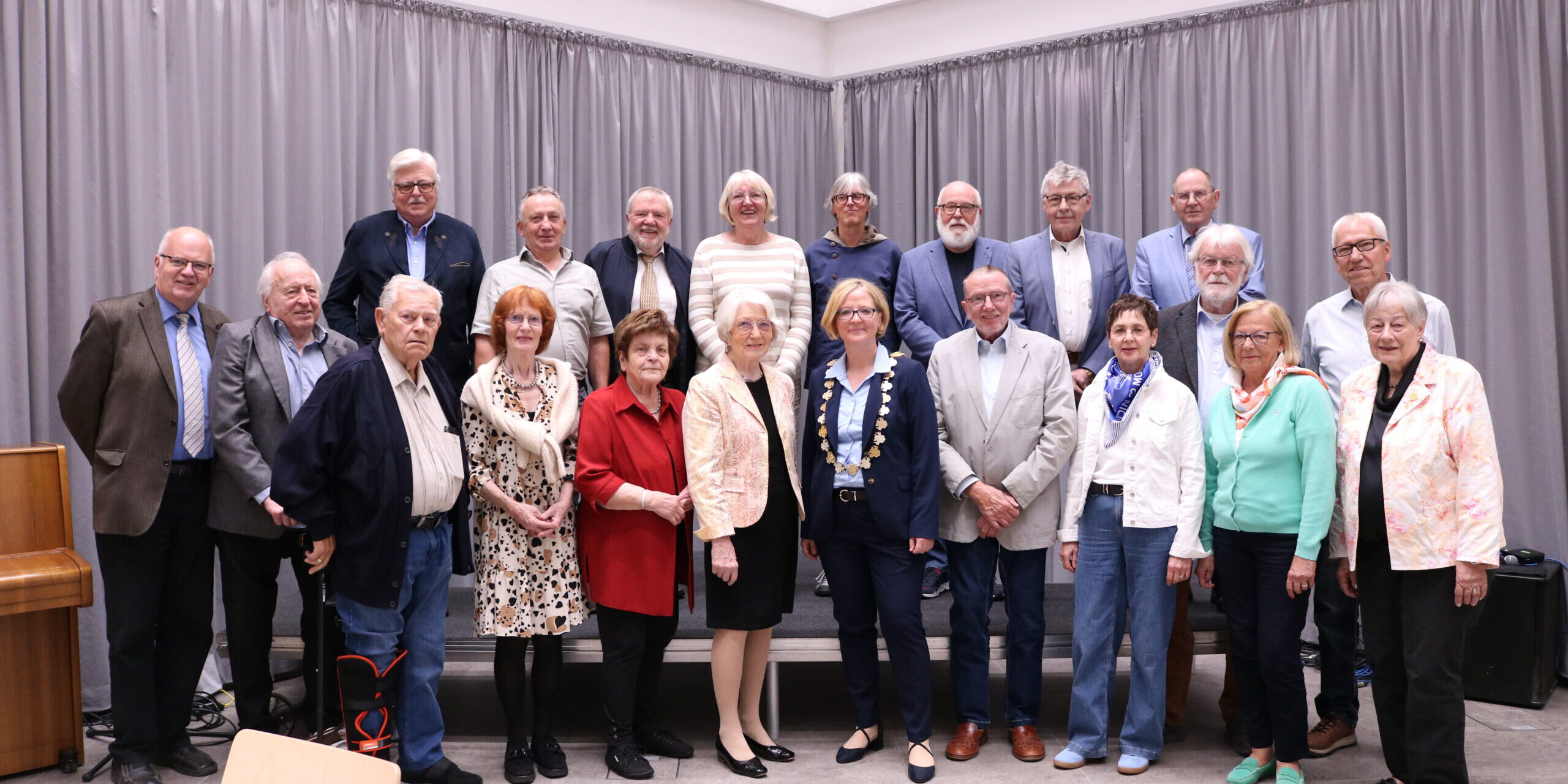 Gruppenfoto einer Ehrungsveranstaltung mit mehr als 20 Personen in einem Saal. Die Teilnehmenden stehen in mehreren Reihen vor einer grauen Vorhangwand. Im Vordergrund befindet sich eine Frau mit Bürgermeisterkette, umgeben von den geehrten Bürgerinnen und Bürgern verschiedener Altersgruppen. Links im Bild ist ein Klavier zu sehen.