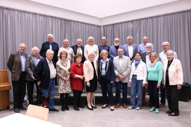 Foto: Stadt Menden/Ehrlich Gruppenfoto einer Ehrungsveranstaltung mit mehr als 20 Personen in einem Saal. Die Teilnehmenden stehen in mehreren Reihen vor einer grauen Vorhangwand. Im Vordergrund befindet sich eine Frau mit Bürgermeisterkette, umgeben von den geehrten Bürgerinnen und Bürgern verschiedener Altersgruppen. Links im Bild ist ein Klavier zu sehen.
