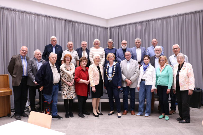 Foto: Stadt Menden/Ehrlich Gruppenfoto einer Ehrungsveranstaltung mit mehr als 20 Personen in einem Saal. Die Teilnehmenden stehen in mehreren Reihen vor einer grauen Vorhangwand. Im Vordergrund befindet sich eine Frau mit Bürgermeisterkette, umgeben von den geehrten Bürgerinnen und Bürgern verschiedener Altersgruppen. Links im Bild ist ein Klavier zu sehen.