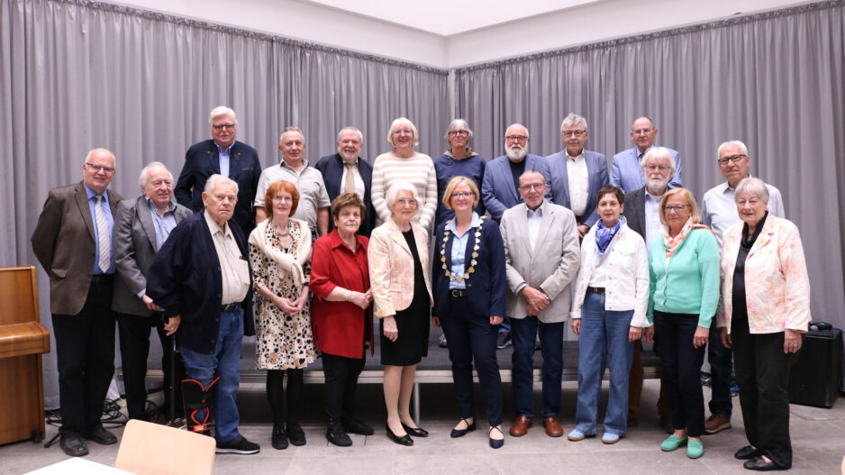 Gruppenfoto einer Ehrungsveranstaltung mit mehr als 20 Personen in einem Saal. Die Teilnehmenden stehen in mehreren Reihen vor einer grauen Vorhangwand. Im Vordergrund befindet sich eine Frau mit Bürgermeisterkette, umgeben von den geehrten Bürgerinnen und Bürgern verschiedener Altersgruppen. Links im Bild ist ein Klavier zu sehen.