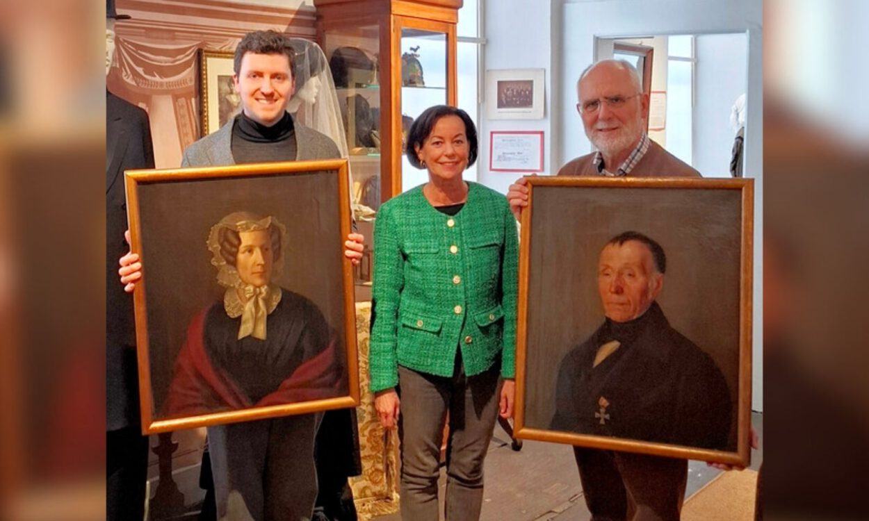 Drei Personen präsentieren in einem Museum zwei historische Porträtgemälde einer Frau und eines Mannes in goldfarbenen Rahmen.