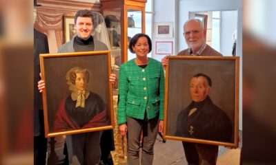 Drei Personen präsentieren in einem Museum zwei historische Porträtgemälde einer Frau und eines Mannes in goldfarbenen Rahmen.