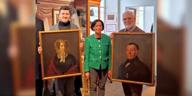 Drei Personen präsentieren in einem Museum zwei historische Porträtgemälde einer Frau und eines Mannes in goldfarbenen Rahmen.