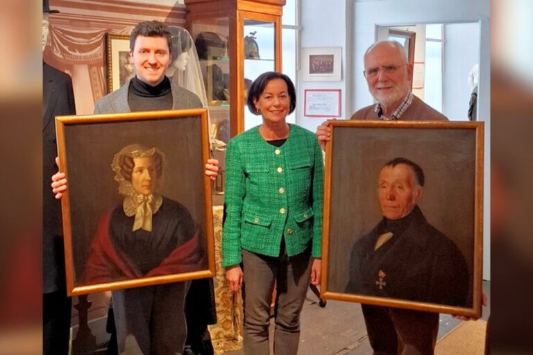 Drei Personen präsentieren in einem Museum zwei historische Porträtgemälde einer Frau und eines Mannes in goldfarbenen Rahmen.