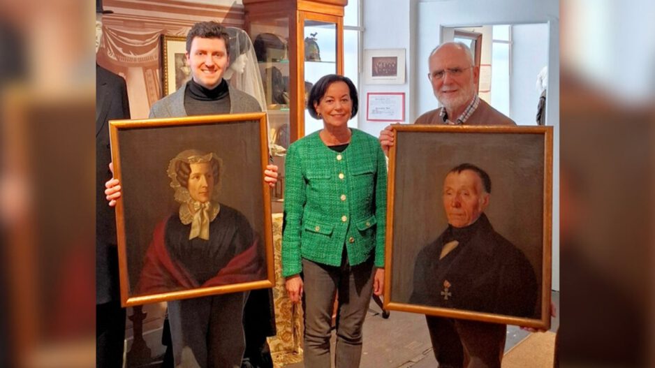 Drei Personen präsentieren in einem Museum zwei historische Porträtgemälde einer Frau und eines Mannes in goldfarbenen Rahmen.