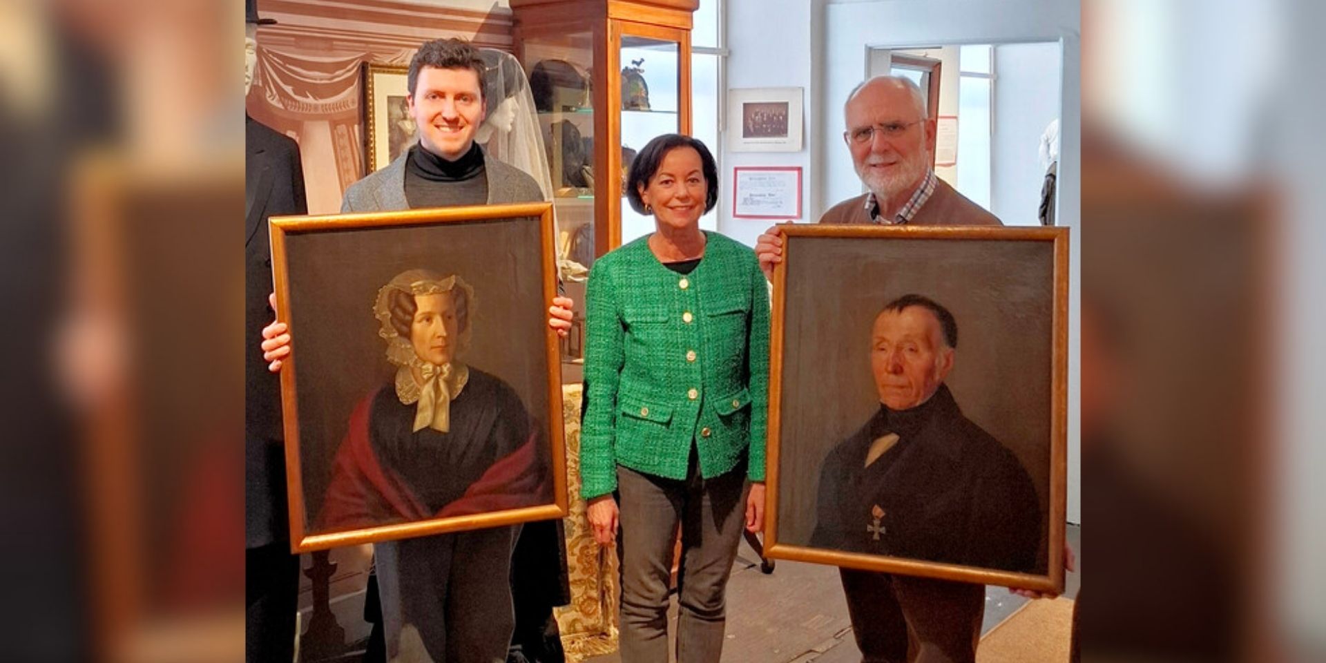 Drei Personen präsentieren in einem Museum zwei historische Porträtgemälde einer Frau und eines Mannes in goldfarbenen Rahmen.