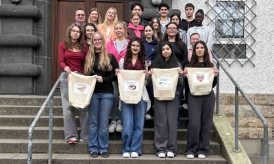 Eine Gruppe von Schülerinnen und Schülern sowie Lehrkräften steht auf den Treppenstufen vor einem Schulgebäude. Vier Jugendliche in der ersten Reihe präsentieren selbst gestaltete Stofftaschen.