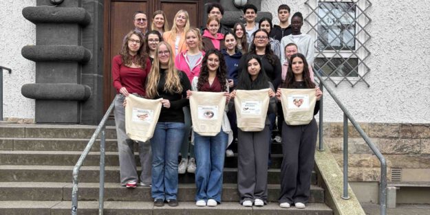 Eine Gruppe von Schülerinnen und Schülern sowie Lehrkräften steht auf den Treppenstufen vor einem Schulgebäude. Vier Jugendliche in der ersten Reihe präsentieren selbst gestaltete Stofftaschen.