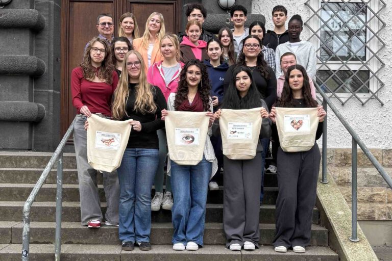 Eine Gruppe von Schülerinnen und Schülern sowie Lehrkräften steht auf den Treppenstufen vor einem Schulgebäude. Vier Jugendliche in der ersten Reihe präsentieren selbst gestaltete Stofftaschen.