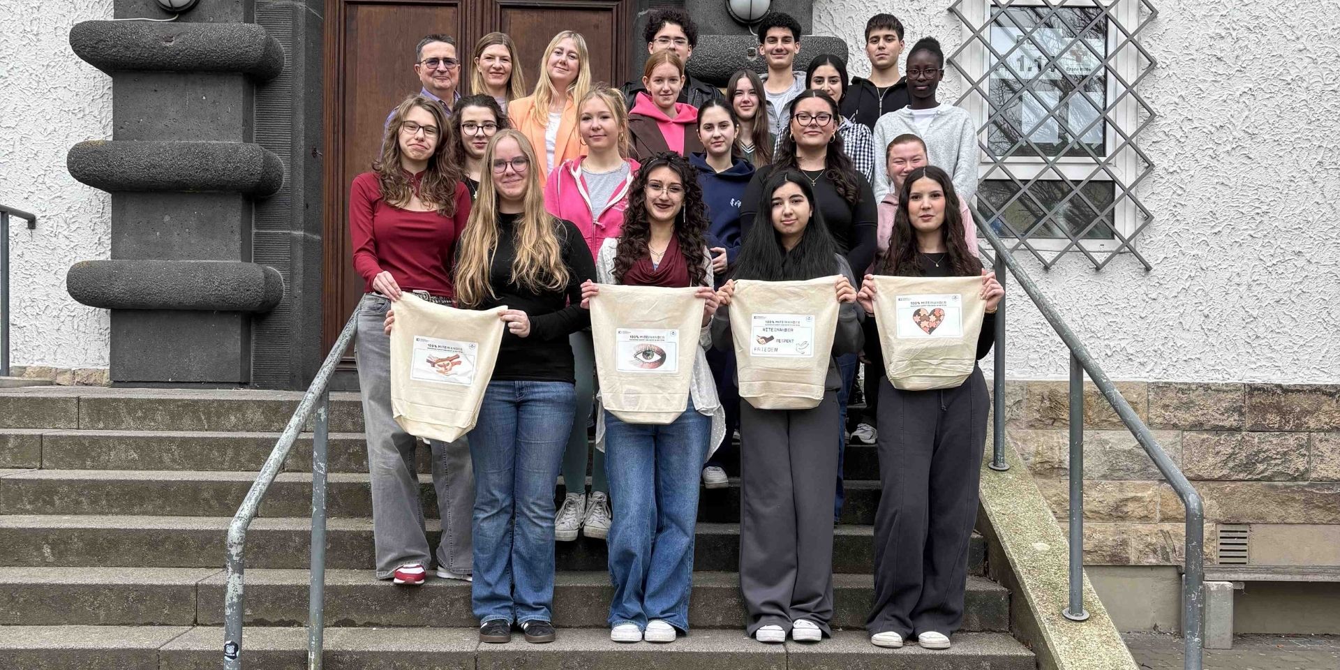 Eine Gruppe von Schülerinnen und Schülern sowie Lehrkräften steht auf den Treppenstufen vor einem Schulgebäude. Vier Jugendliche in der ersten Reihe präsentieren selbst gestaltete Stofftaschen.
