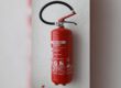 Ein roter Feuerlöscher ist an einer hellen Wand montiert. Das Gerät verfügt über einen schwarzen Löschschlauch und einen Handgriff sowie ein gut sichtbares Etikett mit Bedienhinweisen und technischen Angaben.