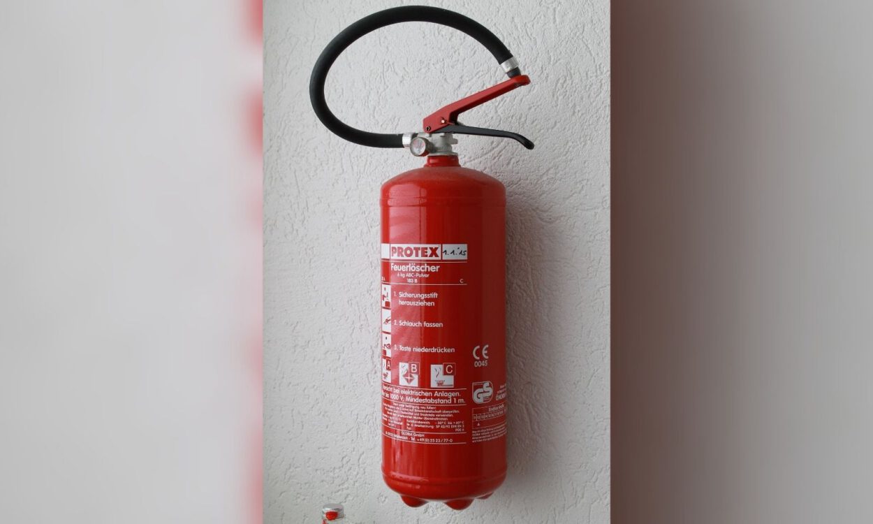 Ein roter Feuerlöscher ist an einer hellen Wand montiert. Das Gerät verfügt über einen schwarzen Löschschlauch und einen Handgriff sowie ein gut sichtbares Etikett mit Bedienhinweisen und technischen Angaben.