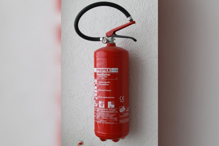 Ein roter Feuerlöscher ist an einer hellen Wand montiert. Das Gerät verfügt über einen schwarzen Löschschlauch und einen Handgriff sowie ein gut sichtbares Etikett mit Bedienhinweisen und technischen Angaben.