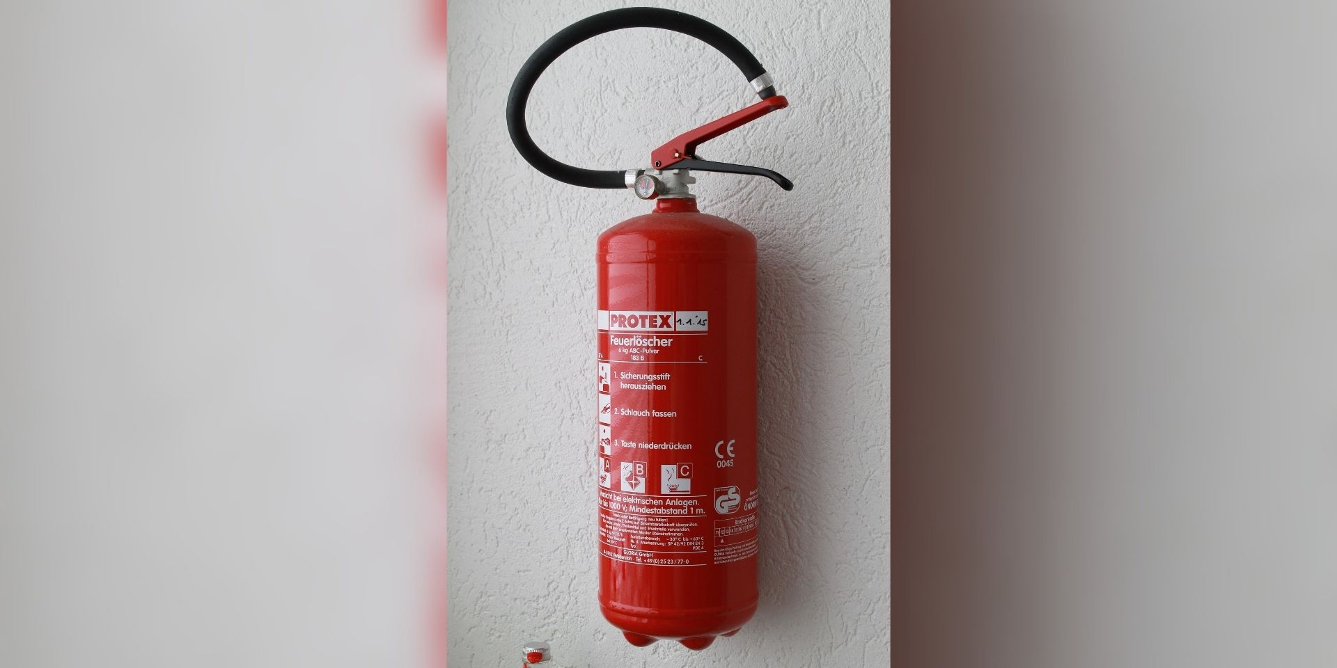 Ein roter Feuerlöscher ist an einer hellen Wand montiert. Das Gerät verfügt über einen schwarzen Löschschlauch und einen Handgriff sowie ein gut sichtbares Etikett mit Bedienhinweisen und technischen Angaben.