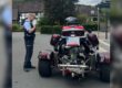 Ein Polizeibeamter steht auf einer Straße neben einem dreirädrigen Motorrad mit breiter Hinterachse. Das Fahrzeug ist von hinten zu sehen und mit Seitenkoffern sowie einem Topcase ausgestattet. Im Hintergrund befinden sich Wohnhäuser und eine Bushaltestelle.