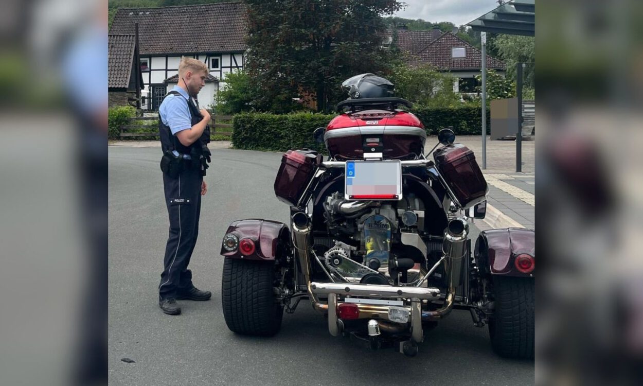 Ein Polizeibeamter steht auf einer Straße neben einem dreirädrigen Motorrad mit breiter Hinterachse. Das Fahrzeug ist von hinten zu sehen und mit Seitenkoffern sowie einem Topcase ausgestattet. Im Hintergrund befinden sich Wohnhäuser und eine Bushaltestelle.