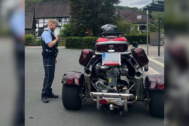 Ein Polizeibeamter steht auf einer Straße neben einem dreirädrigen Motorrad mit breiter Hinterachse. Das Fahrzeug ist von hinten zu sehen und mit Seitenkoffern sowie einem Topcase ausgestattet. Im Hintergrund befinden sich Wohnhäuser und eine Bushaltestelle.